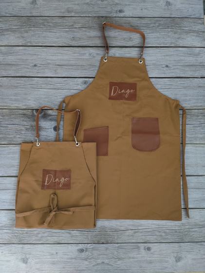Golden brown canvas apron pair with leatherette pockets and script logo patch on grey wood, custom made by RestroCart, New Delhi