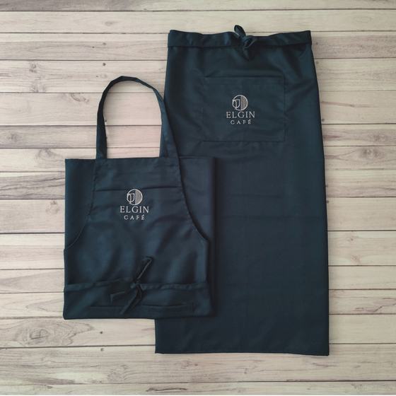 Black bib apron and long black waist apron with grey embroidered logo, laid flat, custom made for Elgin Cafe by RestroCart, New Delhi