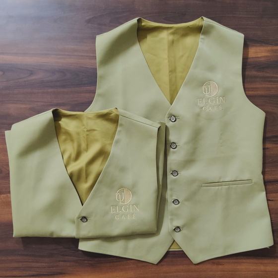 Two olive waistcoats with gold satin lining and tonal embroidered logo, laid on dark wood, custom made for Elgin Cafe by RestroCart, New Delhi