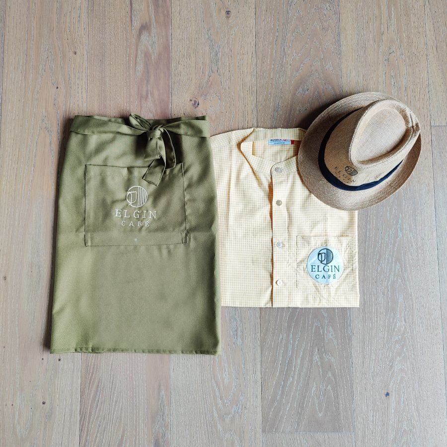 Sage-green apron, cream service shirt with an embroidered logo and a straw fedora, service uniform made for Elgin Cafe, Amritsar, by RestroCart