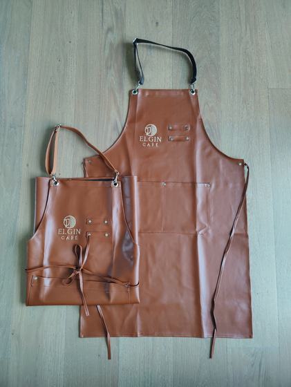 Tan leatherette apron pair with white printed logo and brown ties on a grey floor, custom made for Elgin Cafe by RestroCart, New Delhi