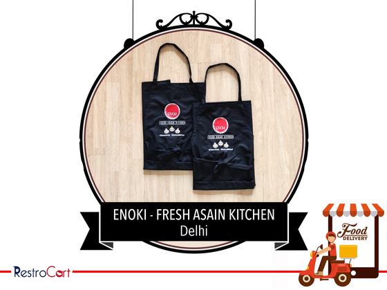 Two black bib aprons with round red logo and white text, laid flat on wood, custom made for Enoki Kitchen by RestroCart, New Delhi