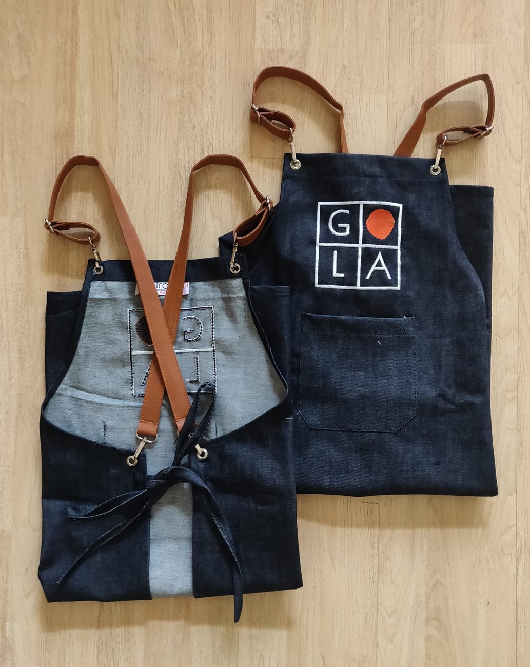 Two indigo denim bib aprons with tan leatherette straps, metal hardware and an embroidered GOLA logo, made for Gola Sizzlers by RestroCart