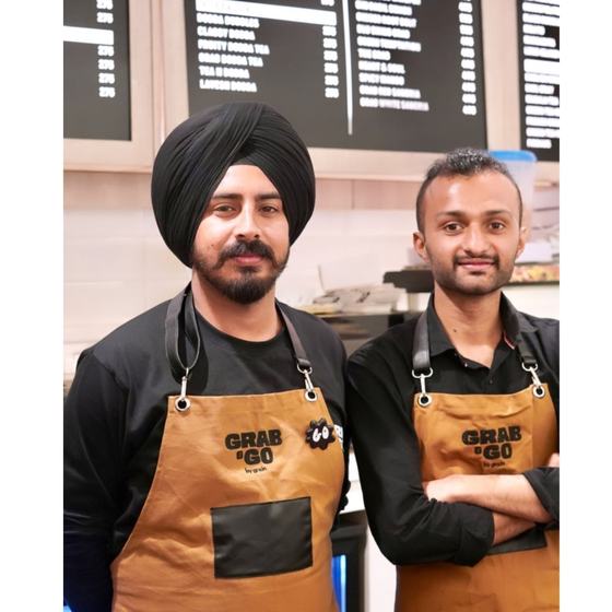 Two counter staff in black shirts wearing tan canvas bib aprons with black straps, metal hooks and black pocket panels, printed with the Grab & Go logo, made by RestroCart