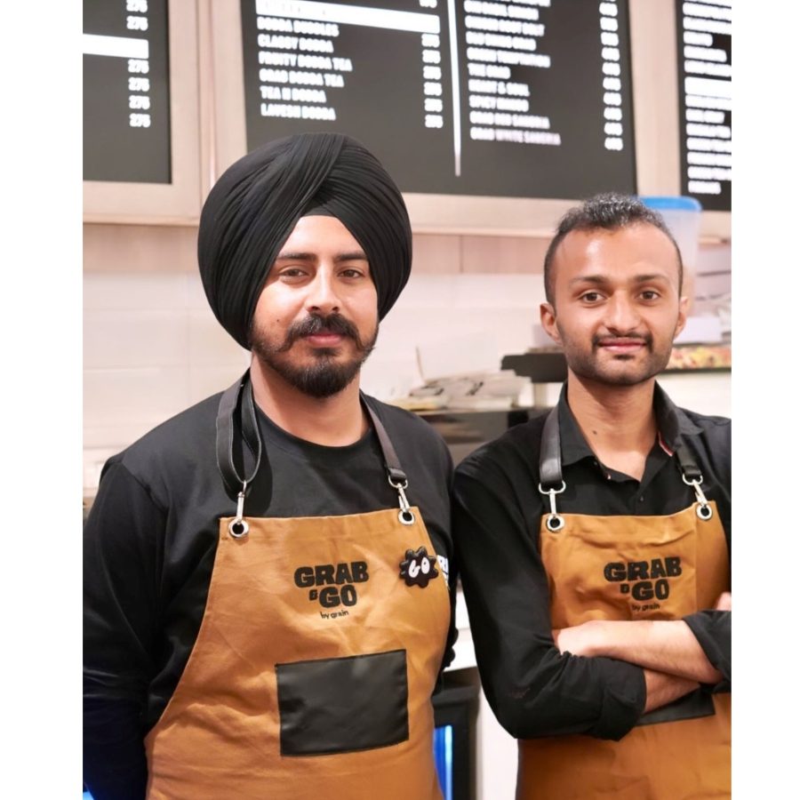 Two counter staff in black shirts wearing tan canvas bib aprons with black straps, metal hooks and black pocket panels, printed with the Grab & Go logo, made by RestroCart