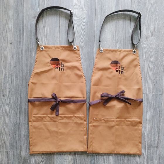 Two tan bib aprons with black leather straps, brown ties and colour-block round logo, custom made for Grain by RestroCart, New Delhi