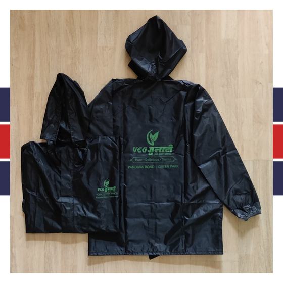 Two black hooded raincoats with green printed Hindi logo on back and chest, laid flat on light wood, custom made for Gulati by RestroCart, New Delhi