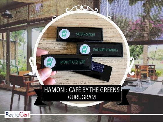 Three black rectangular name badges with green round logo and staff names, one held in hand, custom made for Hamoni Cafe by RestroCart, New Delhi