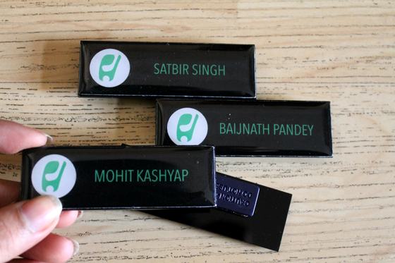 Black glossy name badges with green circular logo and printed staff names, held in hand, custom made for Hamoni Cafe by RestroCart, New Delhi
