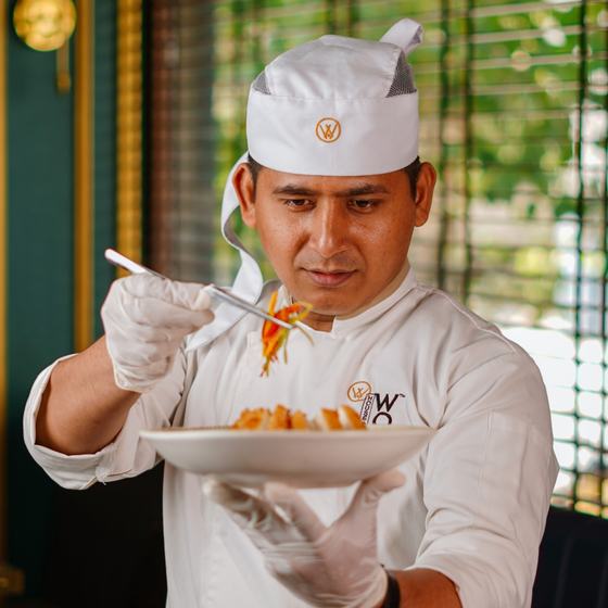Chef plating a dish in a white chef coat and white skull cap with mesh crown, both embroidered with the House of Wok Prive monogram