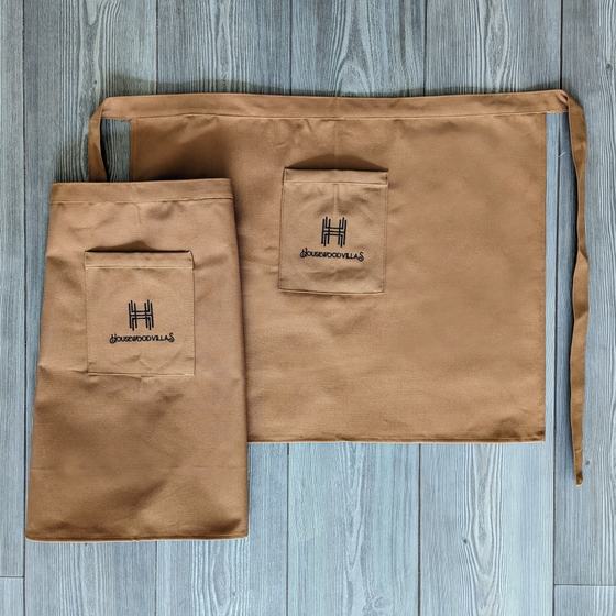 Two tan canvas waist aprons with embroidered logo on front pocket, laid flat, custom made for Housewood Villas by RestroCart, New Delhi