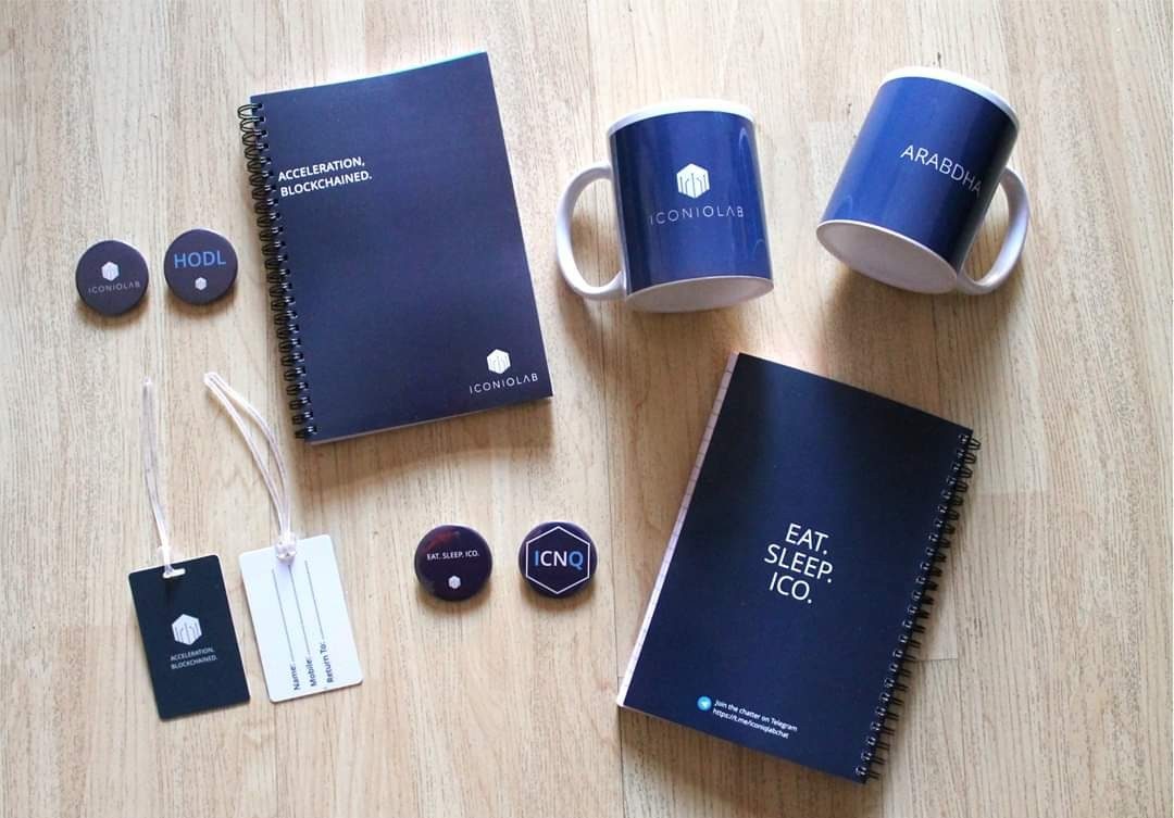 Branded corporate gift kit for IconiqLab laid out flat: navy notebooks, printed mugs, round badges and luggage tags, made by RestroCart