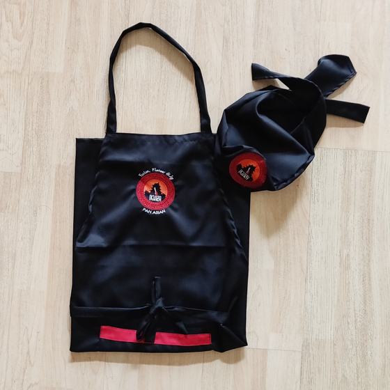 Black bib apron with red hem band and matching skull cap, both with round printed logo, laid flat, custom made for Ikura by RestroCart, New Delhi