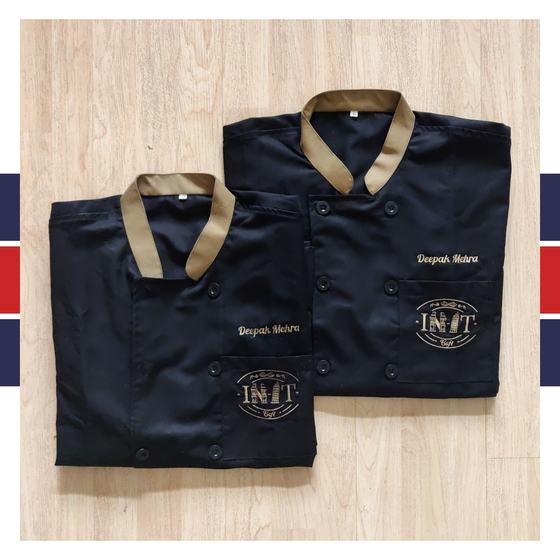 Two folded black chef coats with gold collar, embroidered name and logo on chest pocket, custom made for In It Cafe by RestroCart, New Delhi