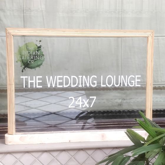 Clear acrylic table sign in light wooden frame with green logo and white event text, custom made for Indian Catering Company by RestroCart, New Delhi