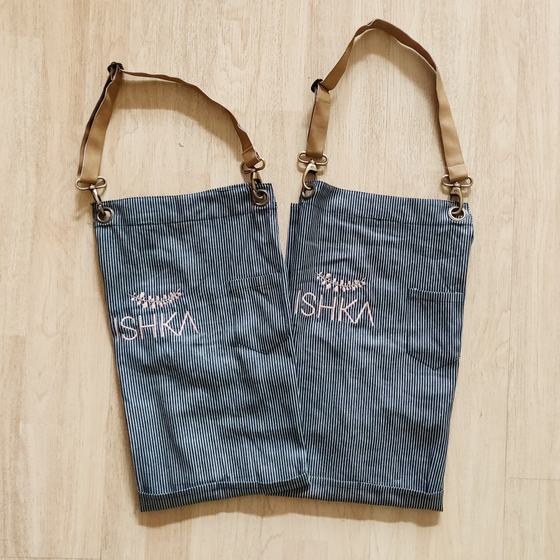 Two grey pinstripe bib aprons with tan straps and metal clips, pink embroidered logo, laid flat, custom made for Ishka Cafe by RestroCart, New Delhi