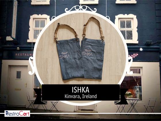 Two grey denim bib aprons with tan leather straps and white printed logo, side by side, custom made for Ishka Cafe by RestroCart, New Delhi