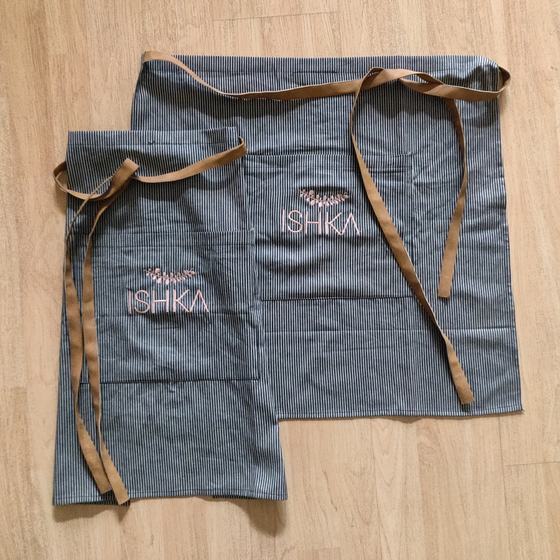 Two grey pinstripe denim waist aprons with tan ties and pink embroidered logo, laid flat, custom made for Ishka Cafe by RestroCart, New Delhi