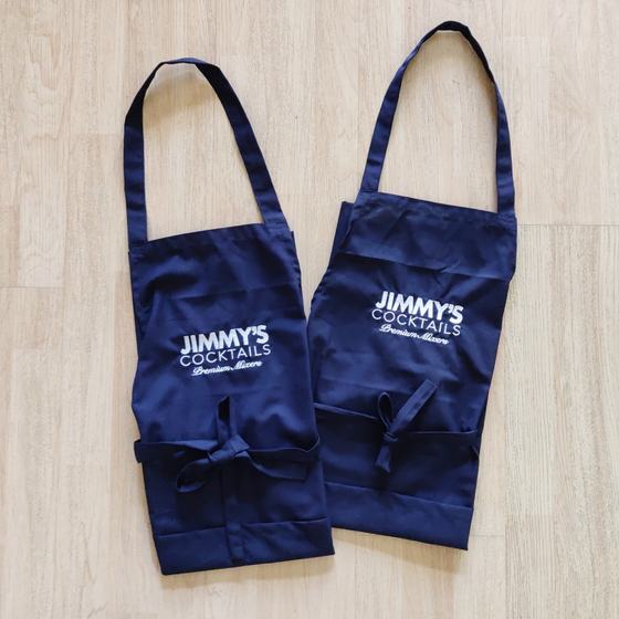 Two navy cotton bib aprons with white printed logo and waist ties, laid flat on wood, custom made for Jimmy's Cocktails by RestroCart, New Delhi