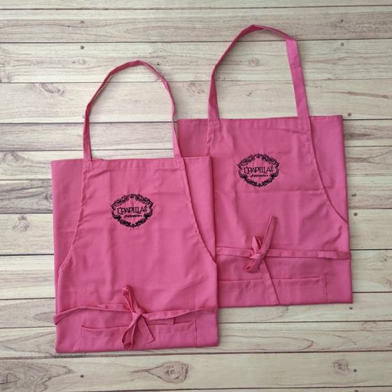 Two pink bib aprons with black embroidered wreath logo and waist ties, laid flat on light wood, custom made for L'Papillae by RestroCart, New Delhi