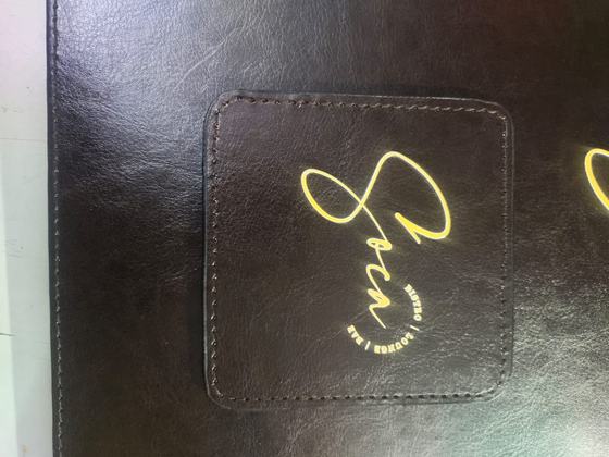 Close-up of a black stitched leatherette coaster with gold foil script logo on a leatherette menu, custom made by RestroCart, New Delhi