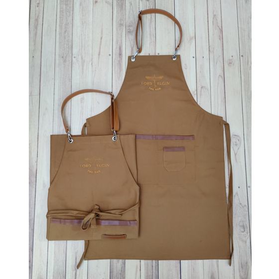 Two tan canvas bib aprons with leather neck straps, chest pocket and printed gold logo, laid flat, custom made for Lord Elgin by RestroCart, New Delhi