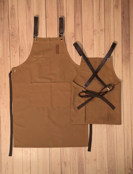 Golden brown canvas apron with leather strap buckles beside a crossback waist apron, custom made by RestroCart, New Delhi