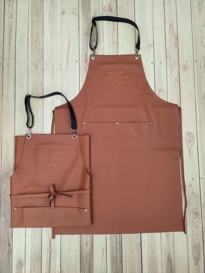 Tan leatherette apron pair with embossed crest logo and rivets, laid on light wood, custom made for Lord Elgin by RestroCart, New Delhi