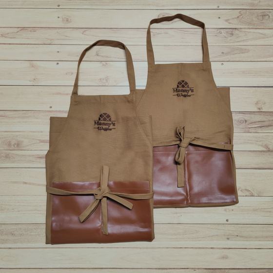 Two tan canvas bib aprons with brown leatherette pocket panel and printed logo, laid flat, custom made for Manny's Waffles by RestroCart, New Delhi