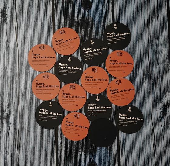 Round MDF coasters in orange and black with printed slogan text, arranged on grey planks, custom made by RestroCart, New Delhi