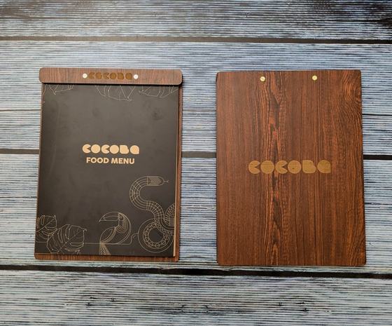 Pair of dark wood MDF clipboard menu boards, one holding a black printed food menu, laid on grey planks, custom made by RestroCart, New Delhi