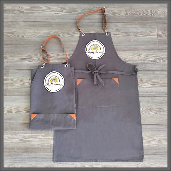 Two grey canvas bib aprons with tan leather straps, orange corner accents and round logo patch, custom made for Melt House by RestroCart, New Delhi