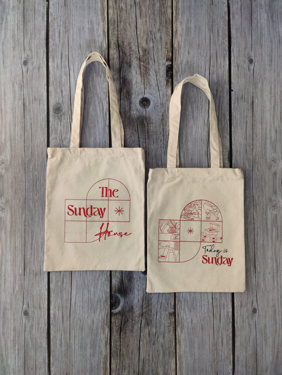 Two natural cotton tote bags printed in red with a cafe logo and line illustrations, laid flat on weathered wooden boards