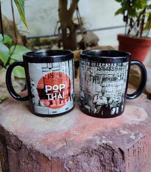 Two black ceramic mugs with black and white street photo print and red circle logo, on a log outdoors, custom made by RestroCart, New Delhi