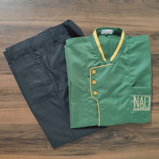 Green chef coat with yellow collar piping and gold buttons, embroidered logo, with grey trousers, custom made for NAO by RestroCart, New Delhi