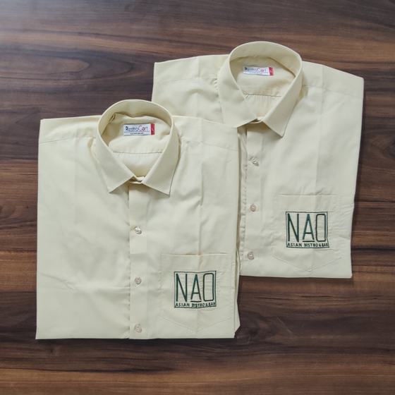 Two cream full sleeve shirts with green embroidered logo on pocket, folded on dark wood, custom made for NAO by RestroCart, New Delhi