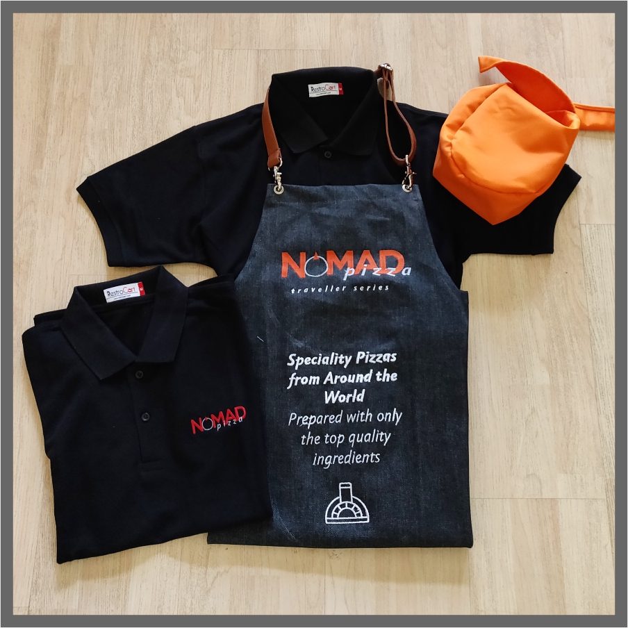 NOMAD Pizza uniform set: black polo t-shirts with embroidered logo, a charcoal denim bib apron with a tan leather strap and printed brand message, and an orange cap