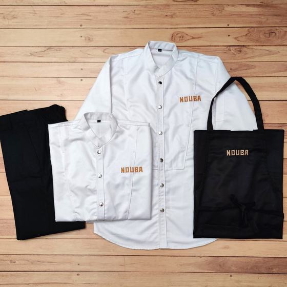 White chef coats with gold embroidered logo, black trousers and black bib apron, laid flat on wood, custom made for Nouba by RestroCart, New Delhi