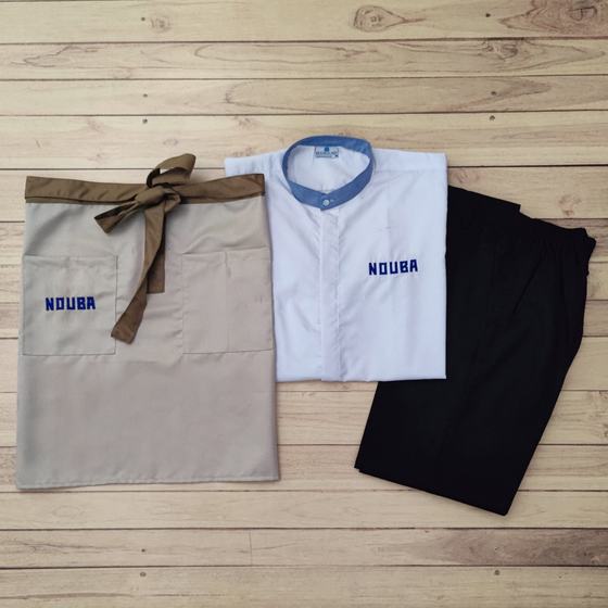 Beige waist apron with brown ties, white shirt with denim collar and black trousers, logo in blue, custom made for Nouba by RestroCart, New Delhi