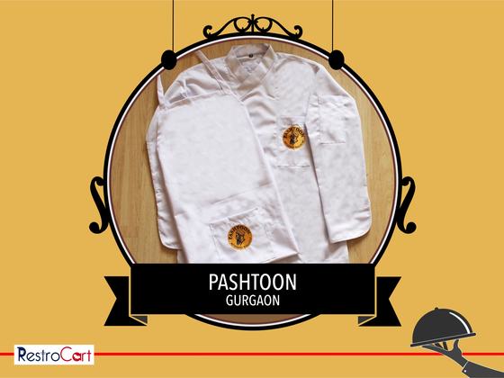 White chef coat and white apron, each with a round orange and black embroidered logo, custom made for Pashtoon by RestroCart, New Delhi