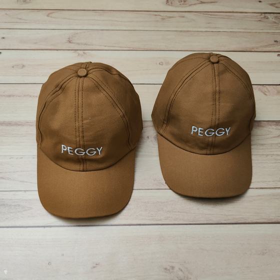Two tan cotton baseball caps with white embroidered wordmark on front, on wooden floor, custom made for Peggy Ice Creams by RestroCart, New Delhi