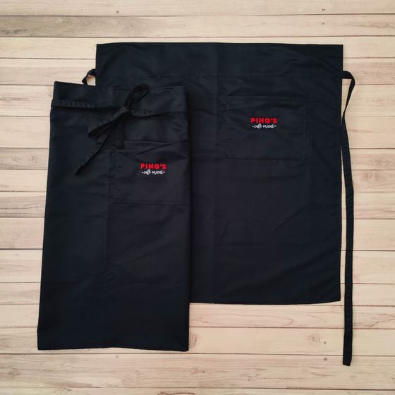 Two black long waist aprons with front pocket and small red embroidered logo, laid flat, custom made for Pings by RestroCart, New Delhi