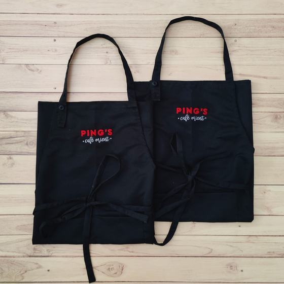 Two black bib aprons with red and white embroidered logo on chest and long waist ties, laid flat, custom made for Pings by RestroCart, New Delhi
