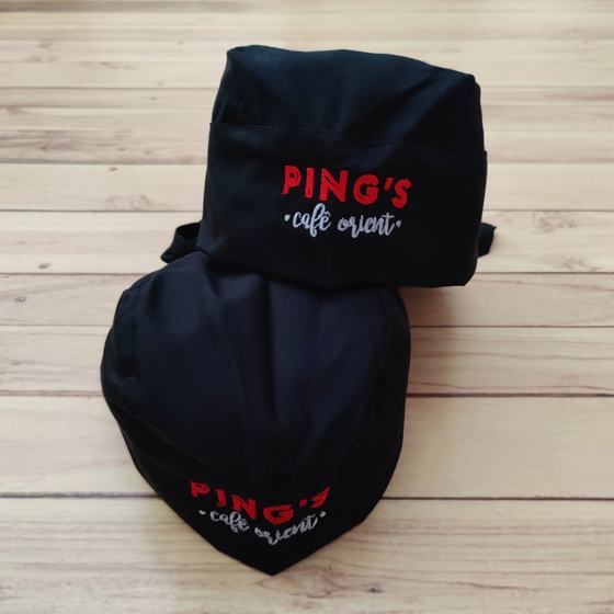 Two black skull caps with red and white embroidered logo, stacked on light wooden floor, custom made for Pings by RestroCart, New Delhi
