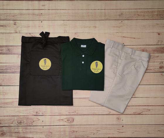 Bottle green polo t-shirt with yellow badge logo, black waist apron and beige trousers, laid flat, custom made by RestroCart, New Delhi