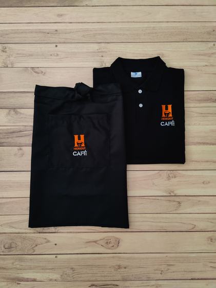 Black polo t-shirt and black waist apron, both with orange and white embroidered cafe logo, on wood, custom made by RestroCart, New Delhi