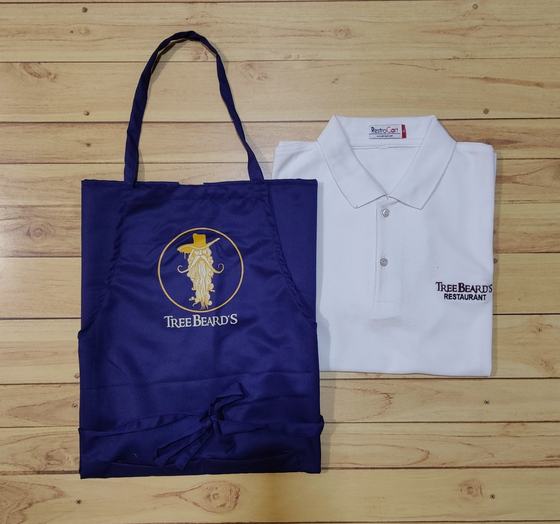 Navy bib apron with gold printed bearded mascot logo beside a white polo t-shirt with text logo, custom made by RestroCart, New Delhi