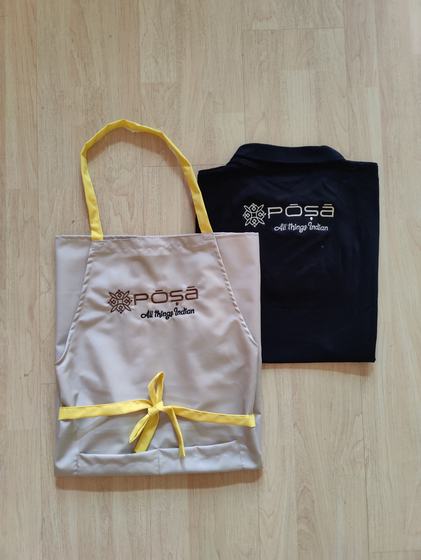 Beige bib apron with yellow straps and brown printed logo beside a folded navy polo t-shirt with logo, custom made by RestroCart, New Delhi
