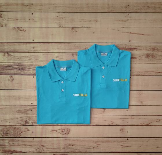 Pair of folded turquoise polo t-shirts with white and yellow printed wordmark on the chest, on wood, custom made by RestroCart, New Delhi