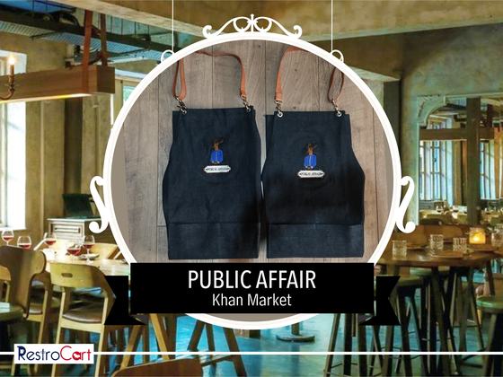 Two dark denim bib aprons with tan leather straps and blue embroidered logo patch, custom made for Public Affair by RestroCart, New Delhi